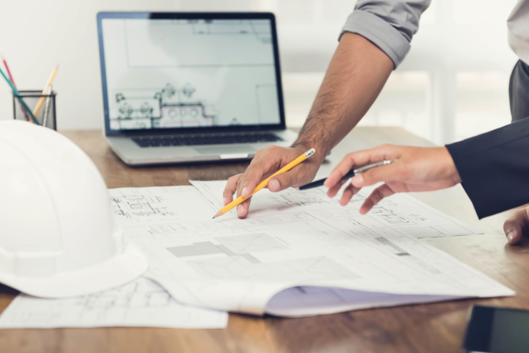 Close-up of two professionals reviewing architectural plans with a pencil and pen in hand, a laptop with design plans on the screen, and a white hard hat in the foreground, indicating a planning or review process in an office environment.