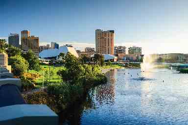 A scenic view of a city skyline with modern buildings by a river with a fountain, greenery along the banks, and clear blue skies.