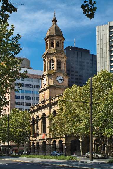  The historic Adelaide Town Hall with its prominent clock tower, nestled among modern buildings and green trees under a blue sky.