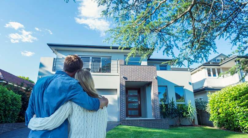  A couple embracing and looking at a modern two-story house with a well-manicured lawn and trees under a clear blue sky.