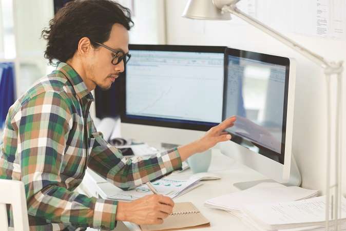 A man with glasses working at a desk with a computer monitor, analyzing data on the screen and taking notes.