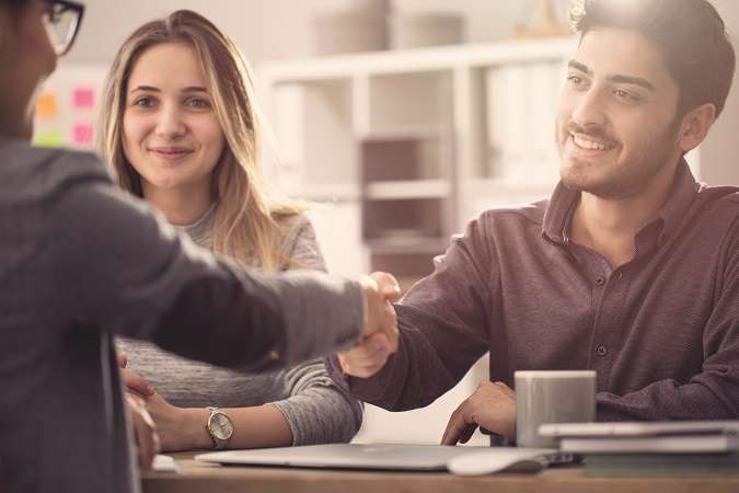 A smiling man and woman shaking hands across a table, likely concluding a meeting or agreement, with another person's hand also visible, suggesting a group interaction.