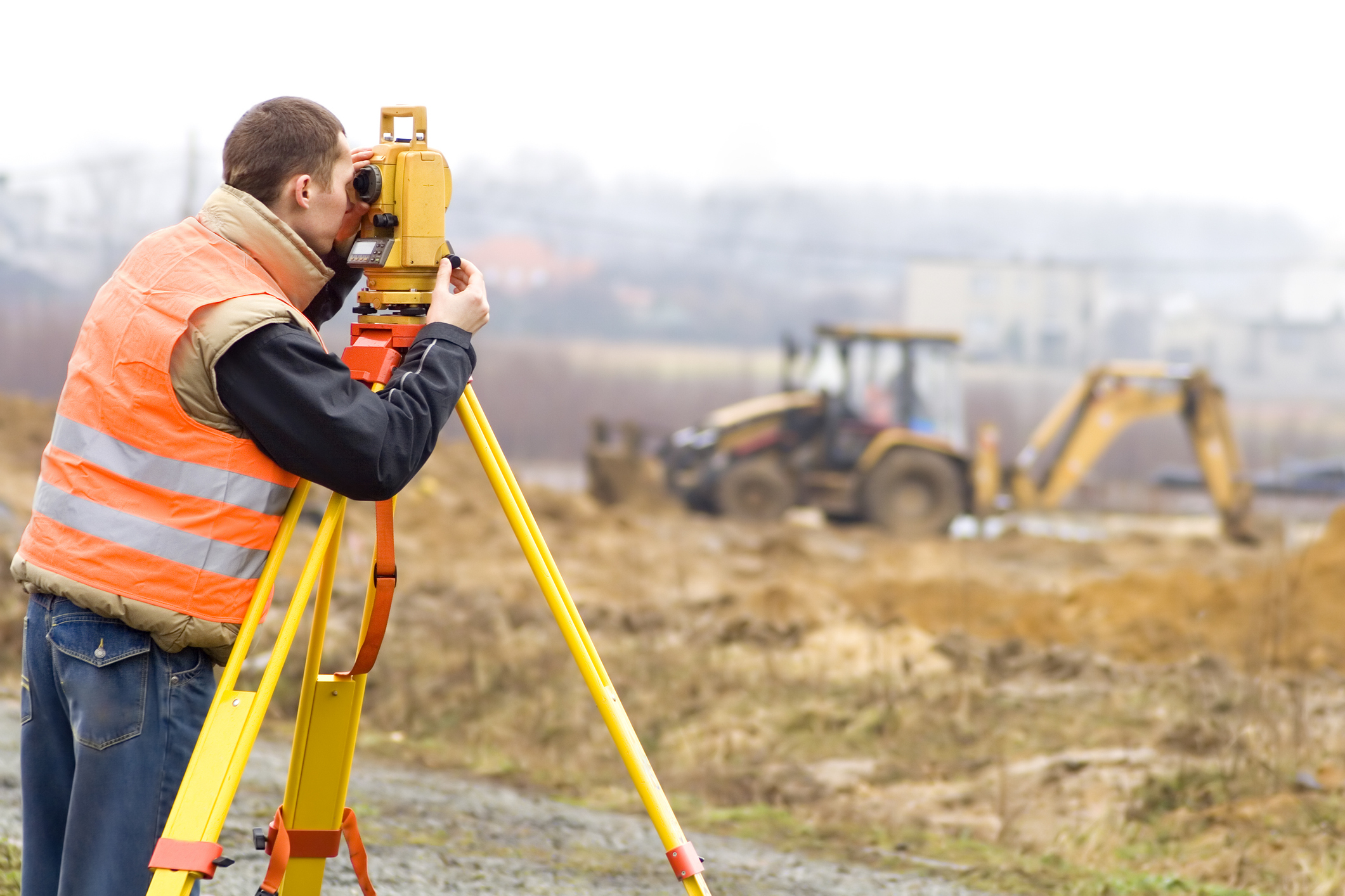 A surveyor using a theodolite on a construction site with heavy machinery in the background.