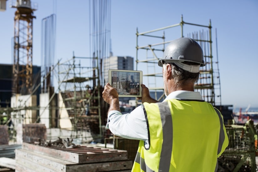  A construction worker in a reflective vest and helmet using a tablet on a construction site with scaffolding and cranes in the background.