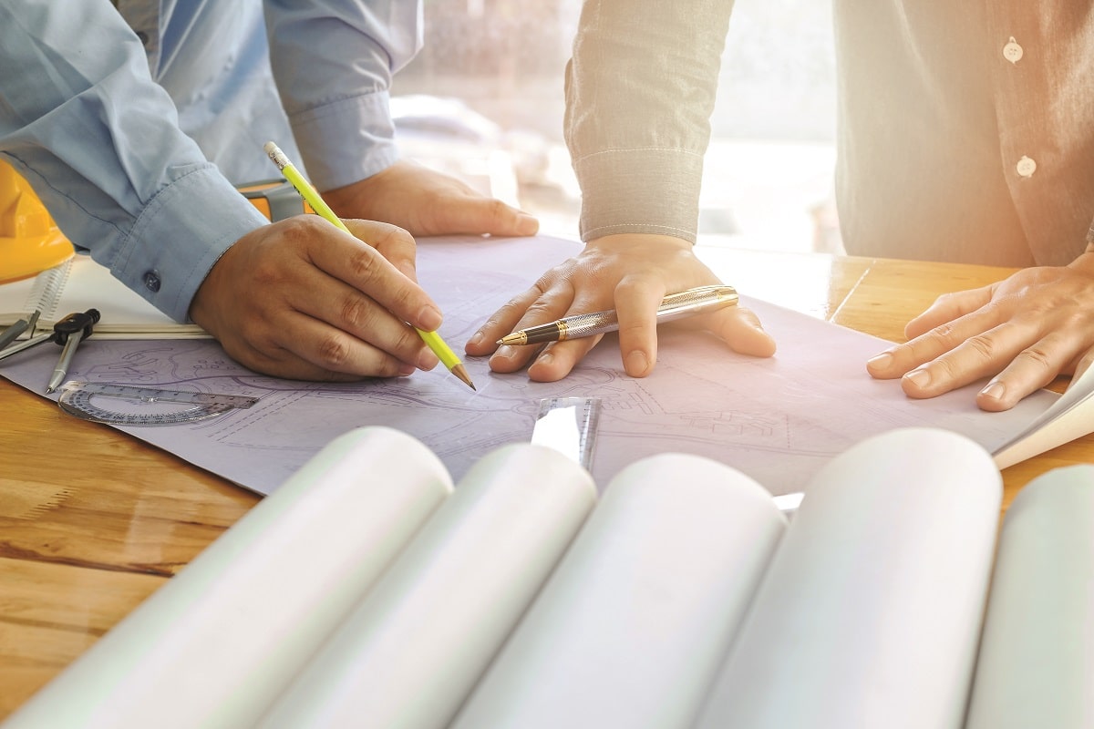 Two people working together on architectural blueprints at a wooden table with drawing tools and a yellow safety helmet visible.