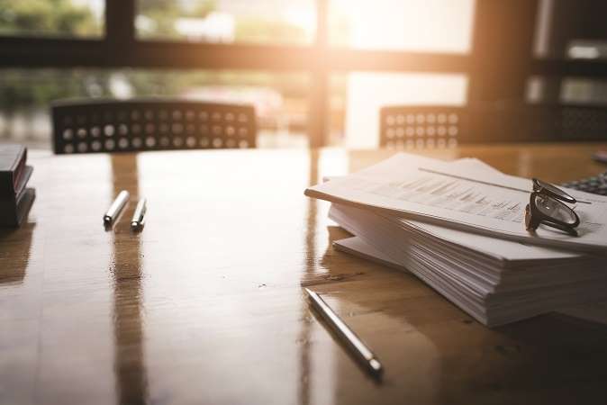  A stack of paperwork with eyeglasses on top and pens nearby on a wooden table, bathed in warm sunlight.
