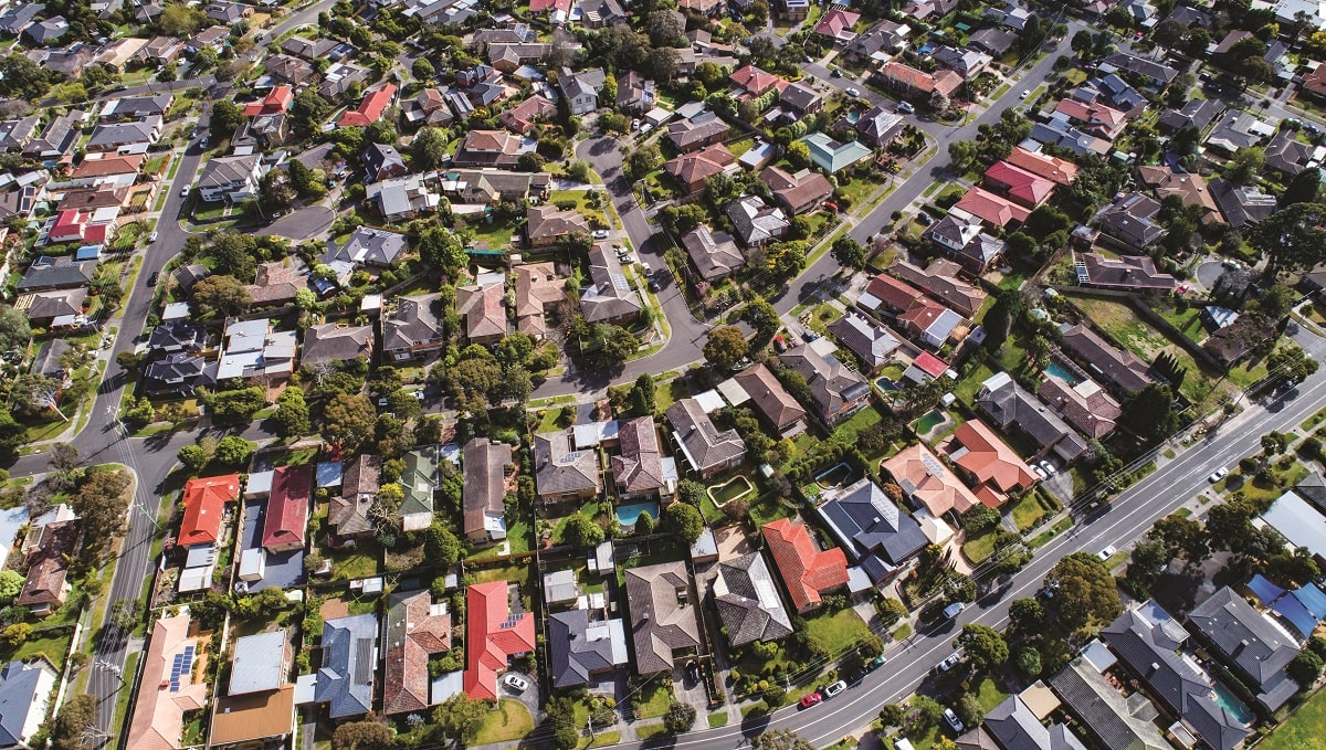 An aerial view of a suburban neighborhood showing a pattern of houses with various roof colors, gardens, and streets.