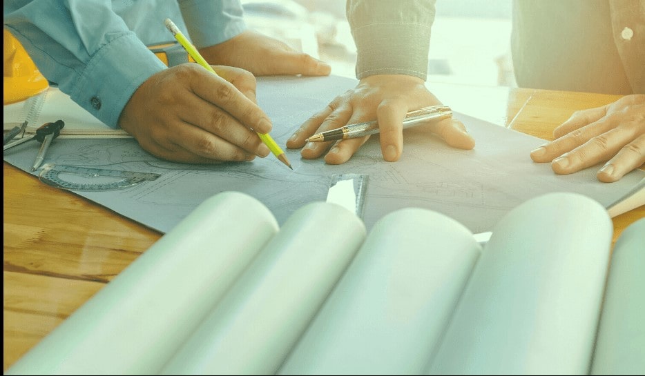 Two people working together on architectural blueprints at a wooden table with drawing tools and a yellow safety helmet visible.